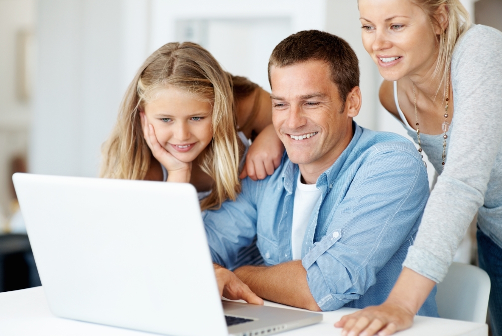 A happy family with a young girl and her parents smiling while looking at a laptop screen together.